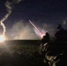 A nighttime military training or combat exercise showing soldiers in the foreground observing multiple pyrotechnic or weapons discharges in the distance. At least two distinct light events are visible: a bright ground-level explosion or flare on the left and a starburst/white phosphorus-type burst in the center. The scene is illuminated primarily by the weapons fire against a dark sky.