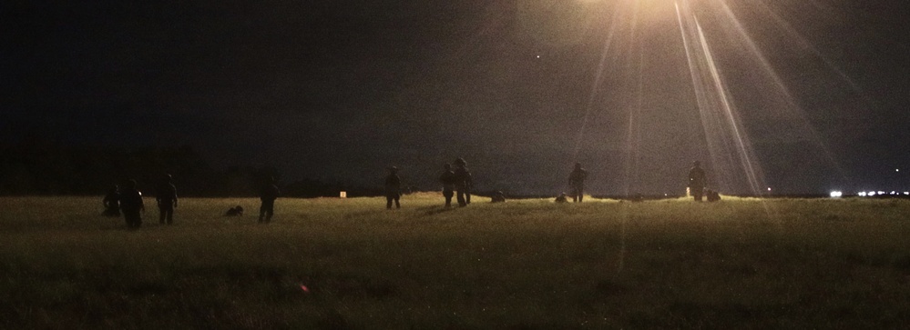 A nighttime outdoor scene showing multiple silhouetted figures spread across an open grassy field. A bright artificial light source in the upper right creates strong lens flare effects, partially illuminating the scene. The figures appear to be engaged in some form of organized ground activity.
