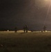 A nighttime outdoor scene showing multiple silhouetted figures spread across an open grassy field. A bright artificial light source in the upper right creates strong lens flare effects, partially illuminating the scene. The figures appear to be engaged in some form of organized ground activity.