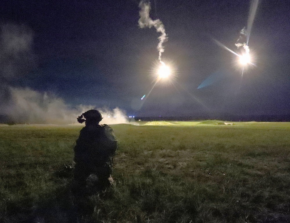 A night-time military field exercise showing a single soldier in full combat gear kneeling on a grass field while illumination flares burn overhead. Two bright aerial flares are visible in the sky, casting light across the terrain and producing smoke trails. The scene is consistent with a nighttime training operation or tactical exercise.