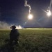 A night-time military field exercise showing a single soldier in full combat gear kneeling on a grass field while illumination flares burn overhead. Two bright aerial flares are visible in the sky, casting light across the terrain and producing smoke trails. The scene is consistent with a nighttime training operation or tactical exercise.
