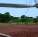 The image is taken through a chain-link fence or goal net, showing a dirt field that appears to be a sports ground. Two individuals in camouflage military uniforms are present on the field along with a dog, suggesting a military working dog (MWD) training exercise. A soccer goal post is visible in the background right, and dense tropical vegetation lines the perimeter.