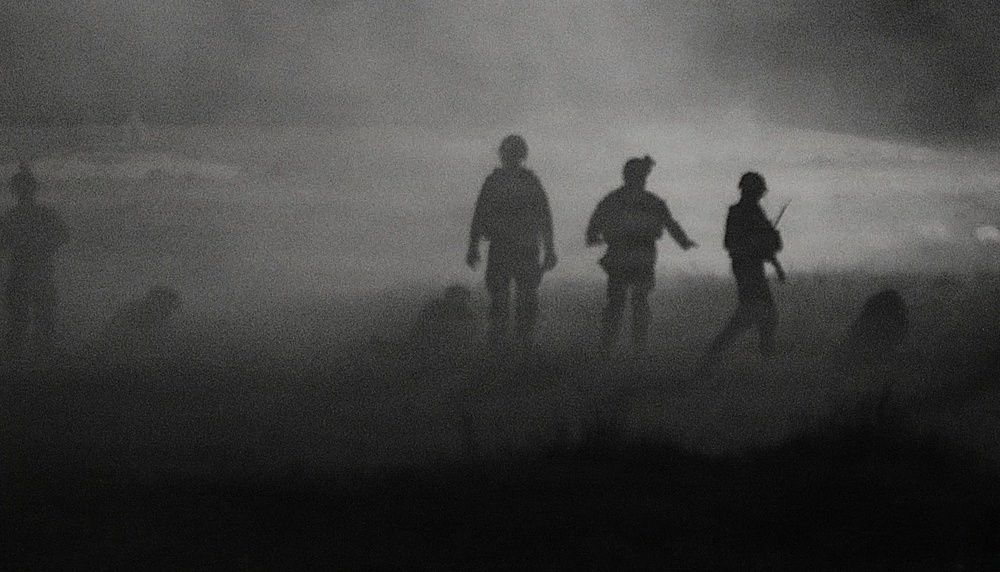 A dark, grainy black-and-white image showing silhouetted figures moving through heavy fog or smoke. At least three central figures are visible in the foreground, with additional shadowy figures on the periphery. The scene has a dramatic, atmospheric quality with a bright light source visible in the background through the haze.