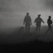 A dark, grainy black-and-white image showing silhouetted figures moving through heavy fog or smoke. At least three central figures are visible in the foreground, with additional shadowy figures on the periphery. The scene has a dramatic, atmospheric quality with a bright light source visible in the background through the haze.