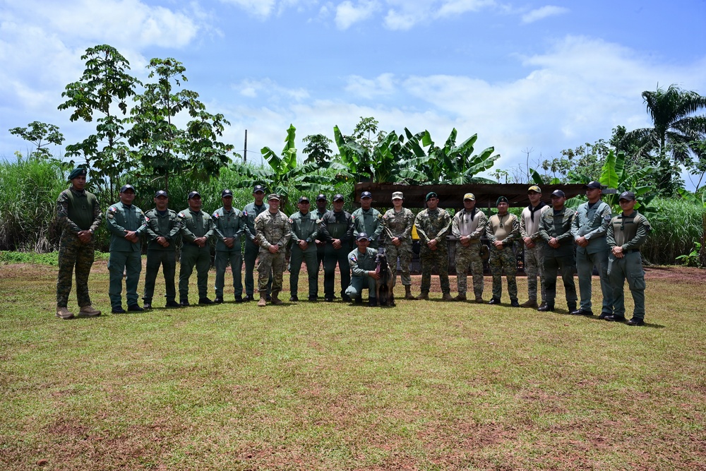 A group of approximately 18-20 military and law enforcement personnel pose for a formal group photograph outdoors in a tropical setting. The group includes individuals in varying uniforms, including green camouflage and tan/multicam patterns, suggesting a multinational or joint training exercise. One individual is kneeling in the front center with a military working dog.
