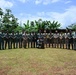 A group of approximately 18-20 military and law enforcement personnel pose for a formal group photograph outdoors in a tropical setting. The group includes individuals in varying uniforms, including green camouflage and tan/multicam patterns, suggesting a multinational or joint training exercise. One individual is kneeling in the front center with a military working dog.