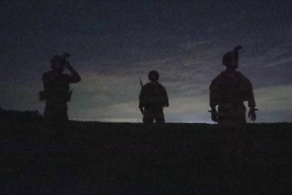 Three silhouetted figures, appearing to be military personnel based on their equipment and helmets, are positioned on elevated ground against a twilight sky. The image is taken at dusk or dawn with minimal ambient light, rendering the subjects almost entirely as dark silhouettes. The scene suggests a tactical observation or patrol scenario.