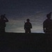 Three silhouetted figures, appearing to be military personnel based on their equipment and helmets, are positioned on elevated ground against a twilight sky. The image is taken at dusk or dawn with minimal ambient light, rendering the subjects almost entirely as dark silhouettes. The scene suggests a tactical observation or patrol scenario.