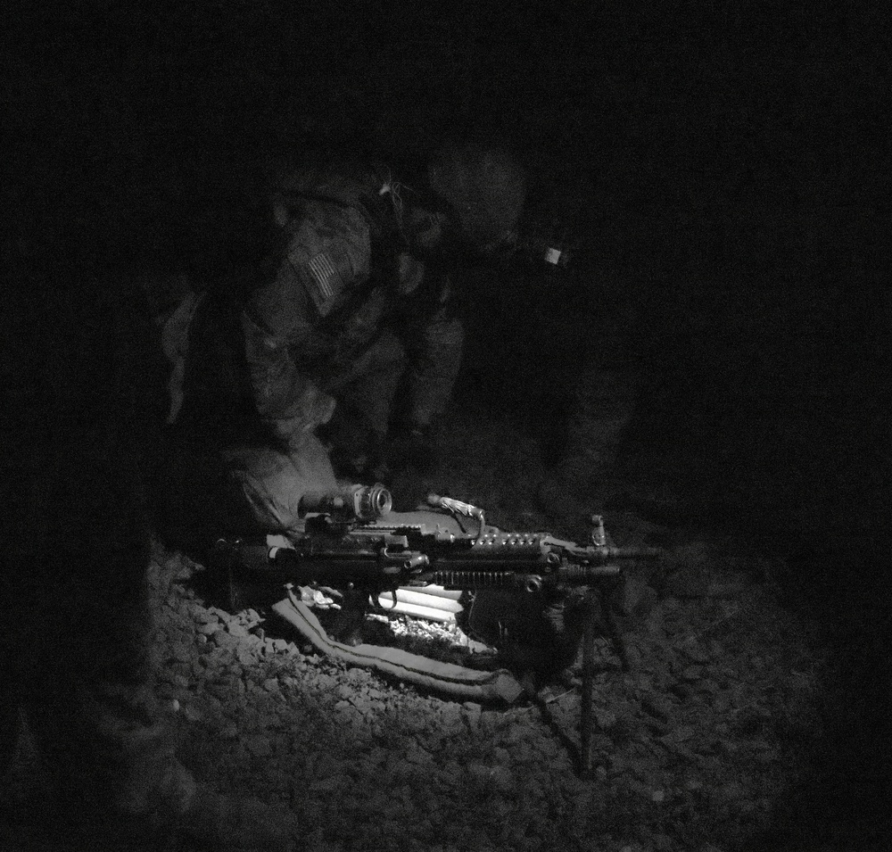 A black-and-white nighttime photograph showing a U.S. soldier in full combat gear crouched over a light machine gun mounted on a bipod. The soldier appears to be performing maintenance, loading, or inspection of the weapon in a low-light field environment. A U.S. flag patch is visible on the soldier's left shoulder.