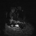 A black-and-white nighttime photograph showing a U.S. soldier in full combat gear crouched over a light machine gun mounted on a bipod. The soldier appears to be performing maintenance, loading, or inspection of the weapon in a low-light field environment. A U.S. flag patch is visible on the soldier's left shoulder.