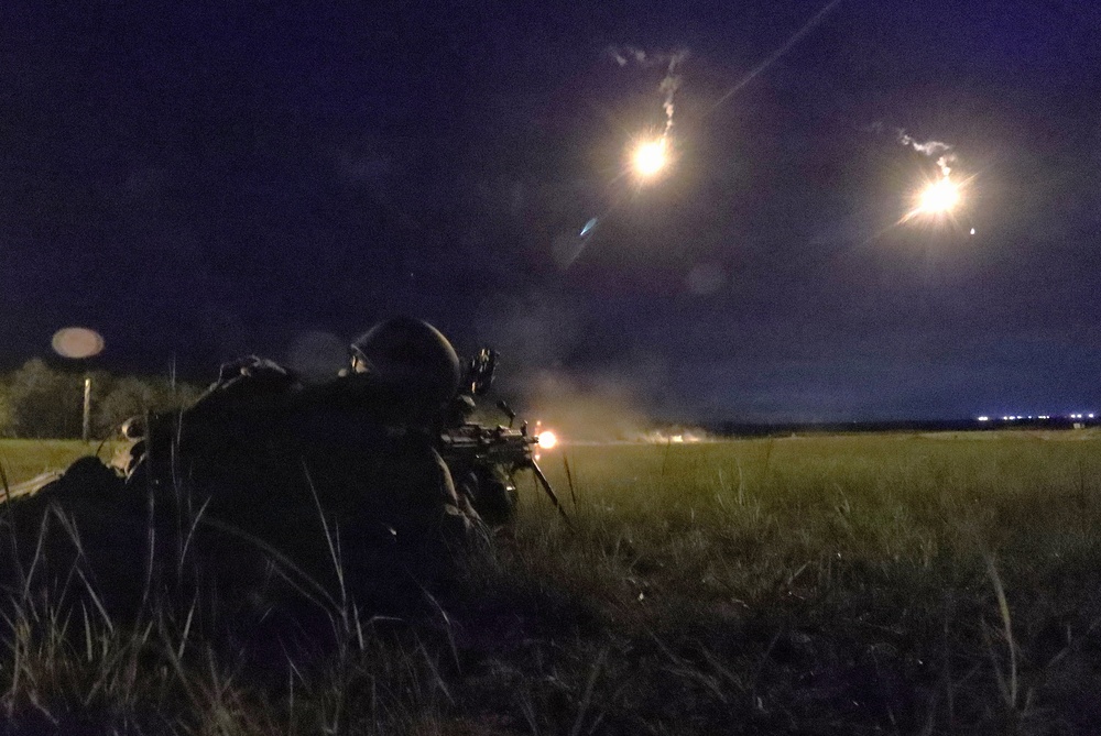 A soldier in full combat gear is in a prone firing position in a grassy field during nighttime operations. The weapon appears to be actively firing, with visible muzzle flash, while illumination flares are deployed overhead to light the area. This appears to be a military training exercise or combat operation conducted under the cover of darkness.