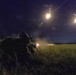 A soldier in full combat gear is in a prone firing position in a grassy field during nighttime operations. The weapon appears to be actively firing, with visible muzzle flash, while illumination flares are deployed overhead to light the area. This appears to be a military training exercise or combat operation conducted under the cover of darkness.