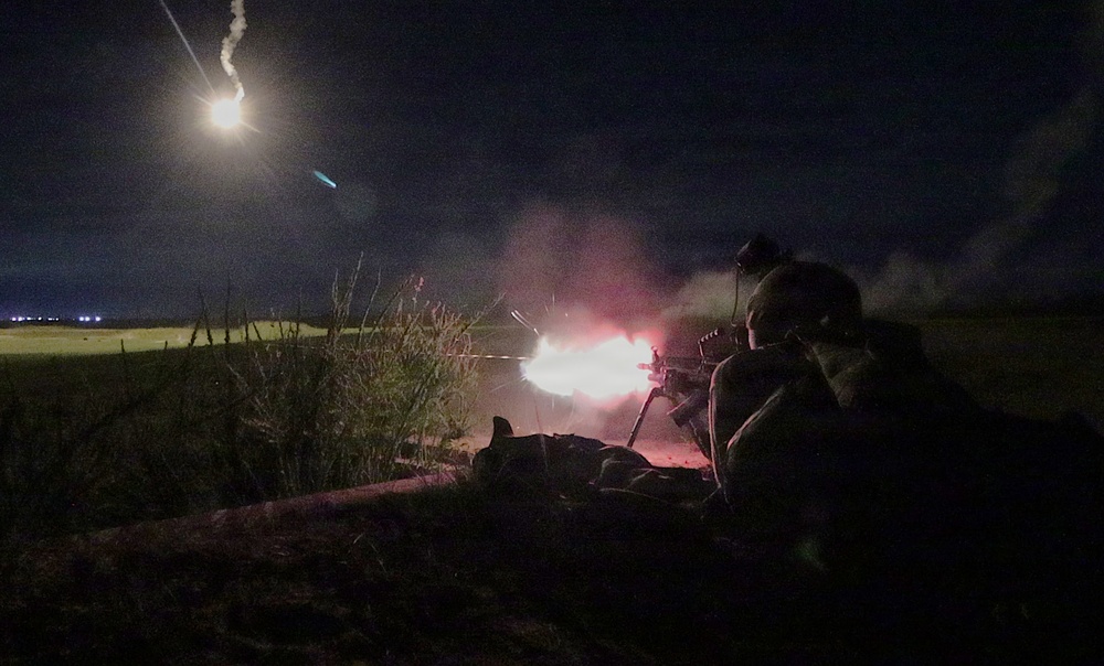 A nighttime military combat or training scene showing a prone soldier firing a light machine gun with a visible muzzle flash. An illumination flare is visible in the upper left sky, casting light over an open field. The scene is characterized by low ambient light with dramatic fire and smoke effects from the weapon discharge.
