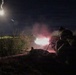 A nighttime military combat or training scene showing a prone soldier firing a light machine gun with a visible muzzle flash. An illumination flare is visible in the upper left sky, casting light over an open field. The scene is characterized by low ambient light with dramatic fire and smoke effects from the weapon discharge.