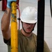 A young male worker wearing a white hard hat and olive green t-shirt is performing maintenance or inspection work on yellow industrial equipment. He is wearing a blue latex glove on his raised hand and has visible tattoos on both arms. The image is accompanied by a caption referencing AGE (Aerospace Ground Equipment) specialist responsibilities.