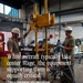 Two military personnel in olive drab uniforms and white hard hats work with ground support equipment inside a maintenance hangar facility. They appear to be inspecting or operating a yellow hydraulic jack or lifting device alongside a tripod stand. The image is accompanied by a quote from 'Luciani' about dispatching ground support equipment to aircraft maintainers on the flightline.