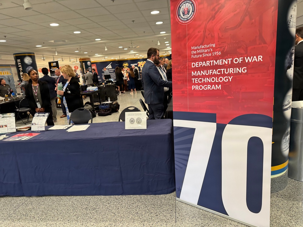 The image shows an exhibition or trade show floor featuring a prominent banner for the 'Department of War Manufacturing Technology Program,' noting it has been 'Manufacturing the Military's Future Since 1956.' A navy-blue draped table is set up in the foreground with informational materials and medallions/coins on display. Multiple attendees and exhibitors are visible in the background at various booths.