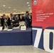 The image shows an exhibition or trade show floor featuring a prominent banner for the 'Department of War Manufacturing Technology Program,' noting it has been 'Manufacturing the Military's Future Since 1956.' A navy-blue draped table is set up in the foreground with informational materials and medallions/coins on display. Multiple attendees and exhibitors are visible in the background at various booths.