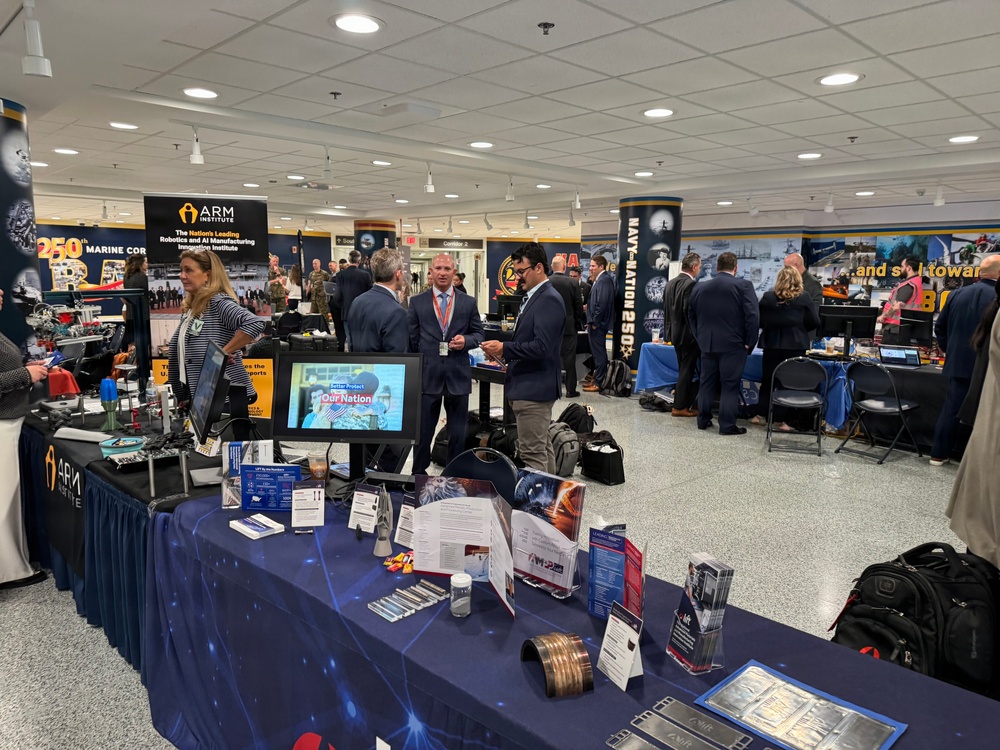 An indoor trade show or exposition featuring multiple vendor booths, including a prominent ARM Institute display promoting robotics and AI manufacturing. The event appears to be military or defense-related, with visible Marine Corps and Navy branding alongside technology exhibitors. Attendees in business attire are networking and reviewing materials at various booths.