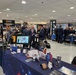 An indoor trade show or exposition featuring multiple vendor booths, including a prominent ARM Institute display promoting robotics and AI manufacturing. The event appears to be military or defense-related, with visible Marine Corps and Navy branding alongside technology exhibitors. Attendees in business attire are networking and reviewing materials at various booths.