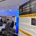This image shows a U.S. Army Manufacturing Technology Program exhibition or trade show display. The prominent banner highlights the Composite Rubber Track (CRT) technology for the Armored Multi-Purpose Vehicle (AMPV), along with a partially visible 'Directed Energy' section. Several attendees in business attire are viewing exhibits in what appears to be an indoor conference or government facility.