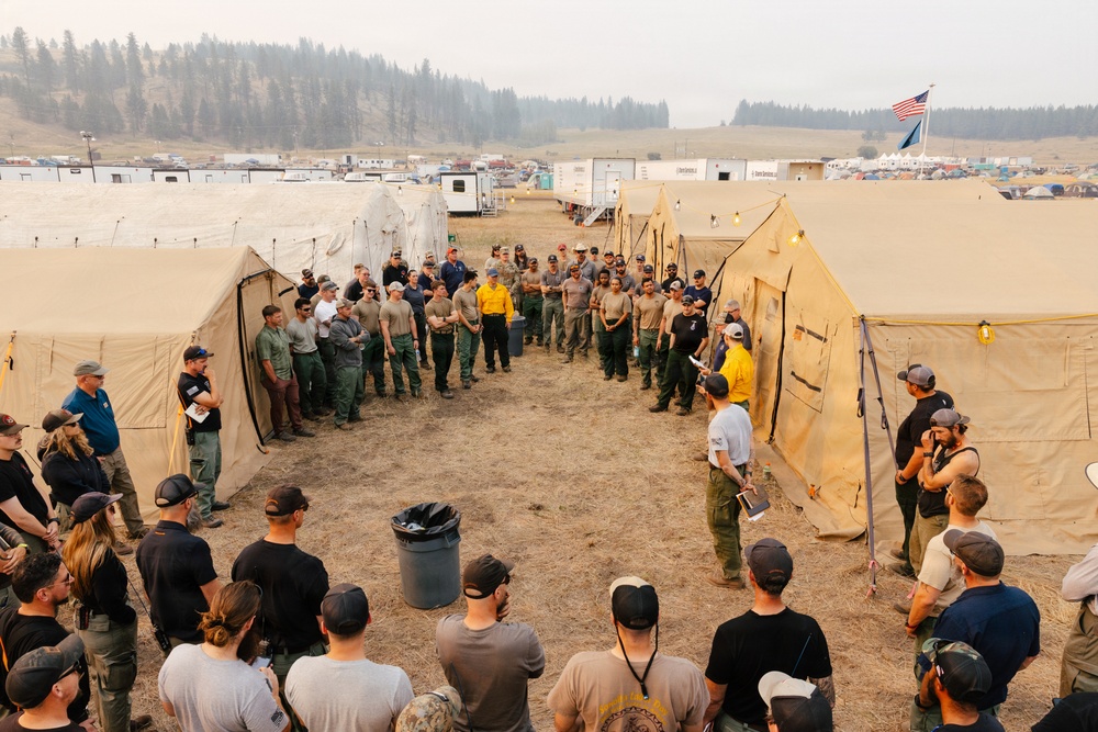 A large group of personnel, likely wildland firefighters, are gathered between rows of tan military-style canvas tents at what appears to be an incident command base camp. Two individuals in yellow high-visibility shirts appear to be conducting a briefing or roll call. The setting includes hazy, smoke-filled skies, an American flag, and numerous support vehicles and trailers in the background, consistent with a wildfire operations base.