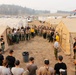 A large group of personnel, likely wildland firefighters, are gathered between rows of tan military-style canvas tents at what appears to be an incident command base camp. Two individuals in yellow high-visibility shirts appear to be conducting a briefing or roll call. The setting includes hazy, smoke-filled skies, an American flag, and numerous support vehicles and trailers in the background, consistent with a wildfire operations base.