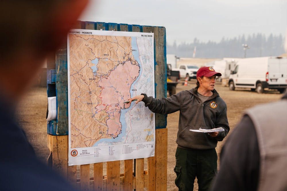 A wildfire incident commander or operations personnel is conducting a briefing using a large fire perimeter map mounted on a wooden pallet stand. The map is labeled 'BRIEFING - MODRITE' with a date of 08/10/2026 and shows fire divisions (DIV S, DIV W, DIV X, DIV A, DIV B, DIV L) with a pink-shaded fire perimeter. The setting appears to be an outdoor incident command post with vehicles visible in the background.