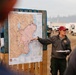 A wildfire incident commander or operations personnel is conducting a briefing using a large fire perimeter map mounted on a wooden pallet stand. The map is labeled 'BRIEFING - MODRITE' with a date of 08/10/2026 and shows fire divisions (DIV S, DIV W, DIV X, DIV A, DIV B, DIV L) with a pink-shaded fire perimeter. The setting appears to be an outdoor incident command post with vehicles visible in the background.