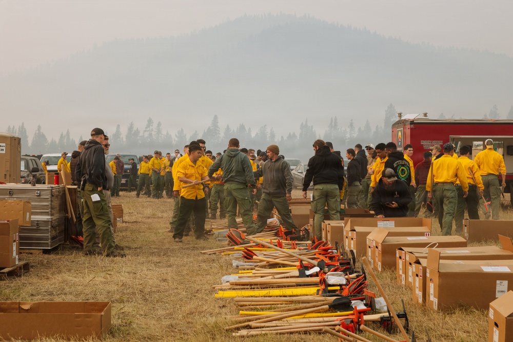 A large group of wildland firefighters are gathered at what appears to be a fire camp or staging area, surrounded by smoke-hazed mountains and conifer forests. Personnel in standard yellow Nomex shirts and green pants are organizing and distributing firefighting hand tools laid out on the ground. The scene suggests a crew mobilization or equipment issue operation prior to deployment.