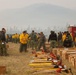 A large group of wildland firefighters are gathered at what appears to be a fire camp or staging area, surrounded by smoke-hazed mountains and conifer forests. Personnel in standard yellow Nomex shirts and green pants are organizing and distributing firefighting hand tools laid out on the ground. The scene suggests a crew mobilization or equipment issue operation prior to deployment.