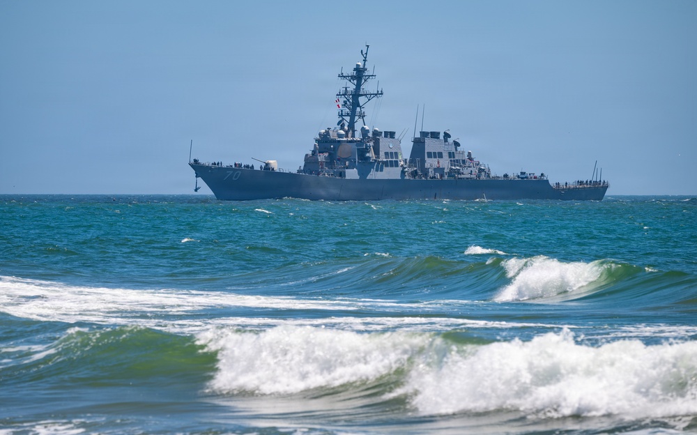 A U.S. Navy Arleigh Burke-class guided-missile destroyer is photographed offshore, viewed from a beach with breaking waves in the foreground. The warship appears to be underway in moderate sea conditions, with personnel visible on deck. The hull number appears to be '70', suggesting this may be USS Hopper or a similarly numbered vessel.