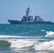 A U.S. Navy Arleigh Burke-class guided-missile destroyer is photographed offshore, viewed from a beach with breaking waves in the foreground. The warship appears to be underway in moderate sea conditions, with personnel visible on deck. The hull number appears to be '70', suggesting this may be USS Hopper or a similarly numbered vessel.