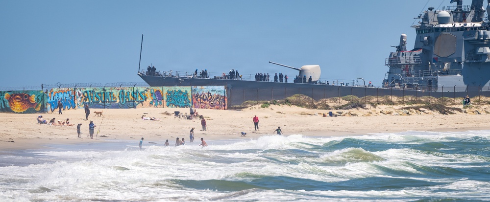 A busy beach scene features civilians swimming and relaxing along a sandy shoreline, with a large naval warship visible in the background docked or anchored near the beach. A concrete wall covered in colorful graffiti murals, including fish artwork and text reading 'Seaside Oasis', separates the beach from the naval vessel area. The contrast between the recreational beach activity and the military ship creates an unusual juxtaposition.