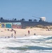 A busy beach scene features civilians swimming and relaxing along a sandy shoreline, with a large naval warship visible in the background docked or anchored near the beach. A concrete wall covered in colorful graffiti murals, including fish artwork and text reading 'Seaside Oasis', separates the beach from the naval vessel area. The contrast between the recreational beach activity and the military ship creates an unusual juxtaposition.