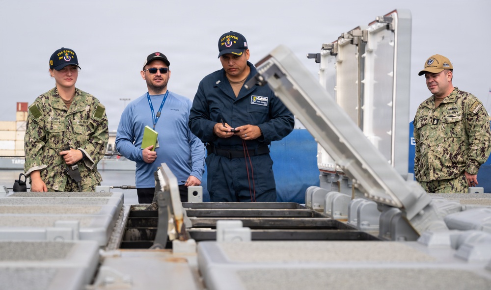 Four individuals, three in U.S. Navy uniforms and one civilian contractor, are gathered on what appears to be the deck of a naval vessel examining equipment. At least two personnel are wearing caps marked 'USS Hopper,' indicating this is an inspection or maintenance activity aboard that ship. The group appears to be reviewing a mechanical hatch or deck access panel with rail/track components.