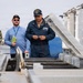 Four individuals, three in U.S. Navy uniforms and one civilian contractor, are gathered on what appears to be the deck of a naval vessel examining equipment. At least two personnel are wearing caps marked 'USS Hopper,' indicating this is an inspection or maintenance activity aboard that ship. The group appears to be reviewing a mechanical hatch or deck access panel with rail/track components.