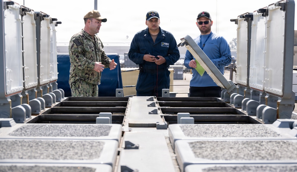 Three individuals are gathered around a piece of military or naval equipment, appearing to conduct an inspection or briefing. One person is wearing U.S. Navy digital camouflage (NWU), another is in a dark blue flight suit or utility uniform, and the third is in civilian attire holding a clipboard or panel. They appear to be on a naval installation or shipyard.