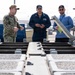 Three individuals are gathered around a piece of military or naval equipment, appearing to conduct an inspection or briefing. One person is wearing U.S. Navy digital camouflage (NWU), another is in a dark blue flight suit or utility uniform, and the third is in civilian attire holding a clipboard or panel. They appear to be on a naval installation or shipyard.