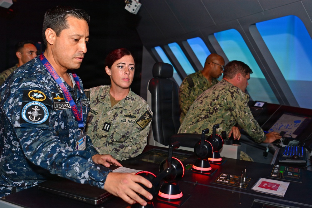 Military personnel from multiple nations are gathered in what appears to be a naval bridge simulator, engaged in training exercises. A male officer wearing a 'República Argentina' navy uniform patch and a female U.S. Navy officer (rank of Lieutenant, surname partially visible as 'MOND') are operating controls at the foreground console. Additional U.S. Navy personnel in NWU (Navy Working Uniform) camouflage are visible operating equipment in the background.