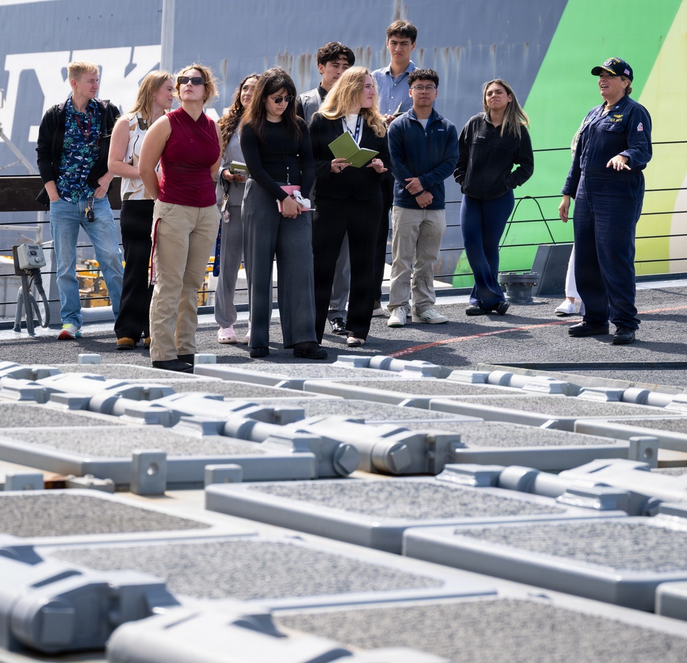 A uniformed naval officer in blue coveralls and a Navy cap is conducting a tour or briefing for a group of approximately 10-12 civilians, likely students, on the deck of a naval vessel. The group is attentively listening as the officer gestures and speaks. In the foreground, gray mechanical deck hardware — likely catapult track components or tie-down fittings — is prominently visible.
