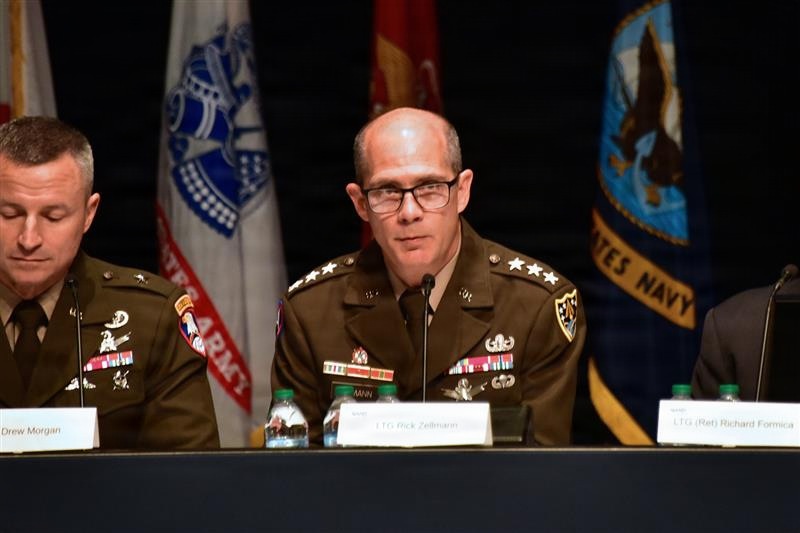 Three military officers are seated at a panel table during what appears to be a formal military conference or symposium. The central figure, identified by a nameplate as LTG Rick Zellmann, is speaking at a microphone while wearing a U.S. Army dress uniform with three-star rank insignia. Military flags including a U.S. Navy flag are visible in the background.