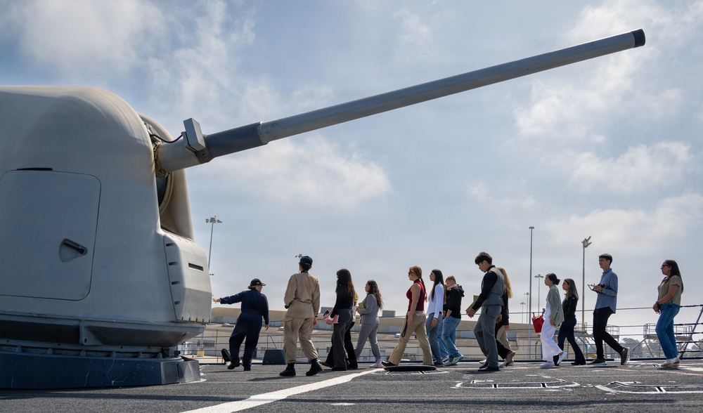 A group of civilians, likely students or visitors, are being given a guided tour on the deck of a naval warship. A uniformed naval officer or crew member is leading the group past a large naval gun turret, which dominates the left foreground of the image. The setting appears to be a naval base or port facility.