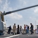 A group of civilians, likely students or visitors, are being given a guided tour on the deck of a naval warship. A uniformed naval officer or crew member is leading the group past a large naval gun turret, which dominates the left foreground of the image. The setting appears to be a naval base or port facility.
