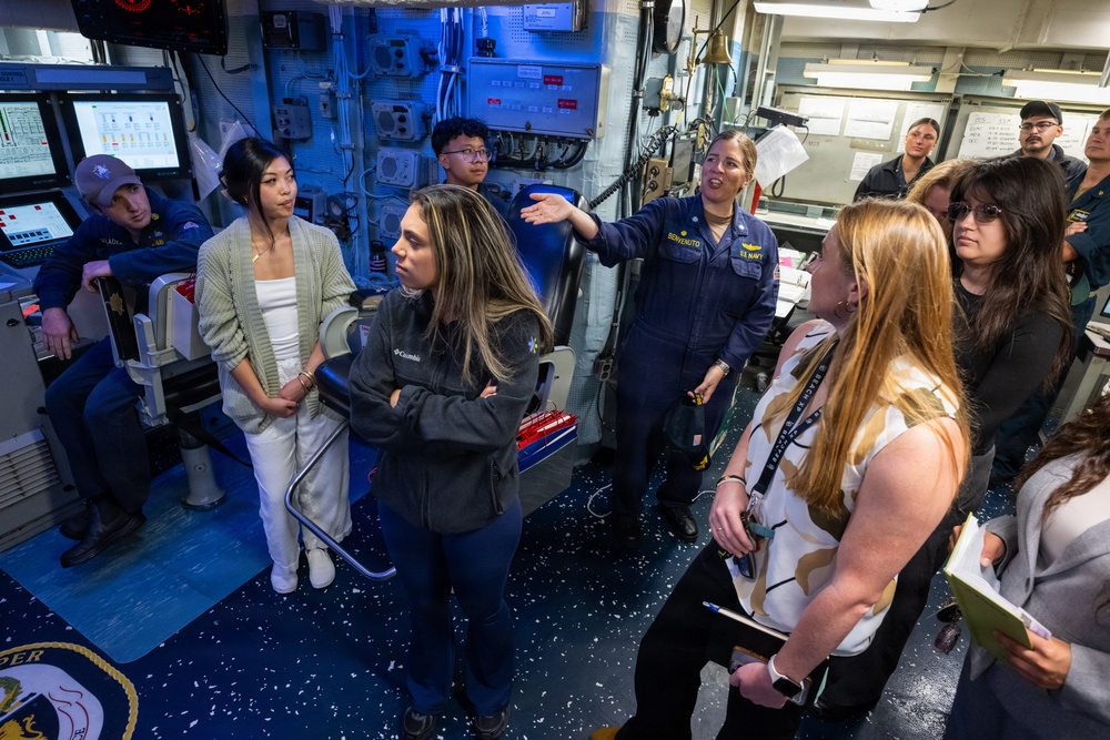 A U.S. Navy sailor in uniform is conducting a tour or briefing for a group of civilians aboard a naval vessel, likely on the bridge or combat information center. The sailor, whose name tag reads 'BENVENUTO,' is gesturing and explaining equipment to the attentive group. Several civilians are taking notes, suggesting this may be an official visit, educational tour, or community outreach event.