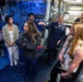 A U.S. Navy sailor in uniform is conducting a tour or briefing for a group of civilians aboard a naval vessel, likely on the bridge or combat information center. The sailor, whose name tag reads 'BENVENUTO,' is gesturing and explaining equipment to the attentive group. Several civilians are taking notes, suggesting this may be an official visit, educational tour, or community outreach event.
