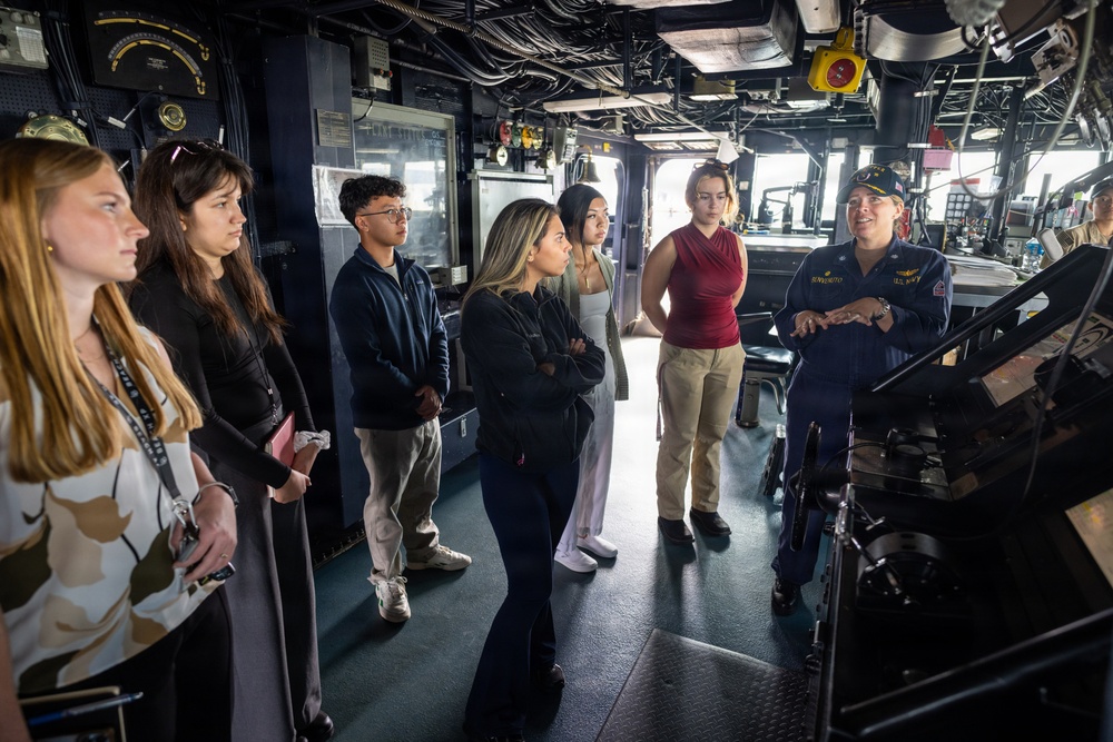 A uniformed U.S. Navy officer is briefing a group of approximately seven civilian visitors on the bridge/pilothouse of a naval vessel. The officer, wearing a blue Navy uniform and cover (cap), appears to be conducting an official tour or educational outreach event. The visitors, a diverse group of young adults, are attentively listening while standing among navigational equipment and ship controls.