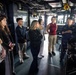 A uniformed U.S. Navy officer is briefing a group of approximately seven civilian visitors on the bridge/pilothouse of a naval vessel. The officer, wearing a blue Navy uniform and cover (cap), appears to be conducting an official tour or educational outreach event. The visitors, a diverse group of young adults, are attentively listening while standing among navigational equipment and ship controls.