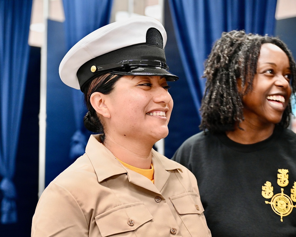Two women are smiling and appear to be at a military ceremony or event. The woman in the foreground is wearing a U.S. Navy enlisted uniform with a white combination cover (cap) and tan service uniform shirt. The woman in the background is wearing a black t-shirt with what appears to be a Navy chief petty officer insignia embroidered on it.