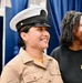 Two women are smiling and appear to be at a military ceremony or event. The woman in the foreground is wearing a U.S. Navy enlisted uniform with a white combination cover (cap) and tan service uniform shirt. The woman in the background is wearing a black t-shirt with what appears to be a Navy chief petty officer insignia embroidered on it.